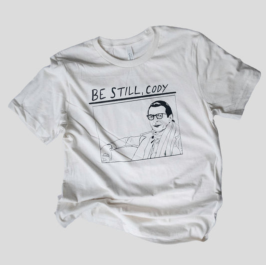 white screen printed t shirt with "be still cody" and a drawing of jeff goldnum as alistair hennessy in the line art style of the cover of sonic youth's album goo